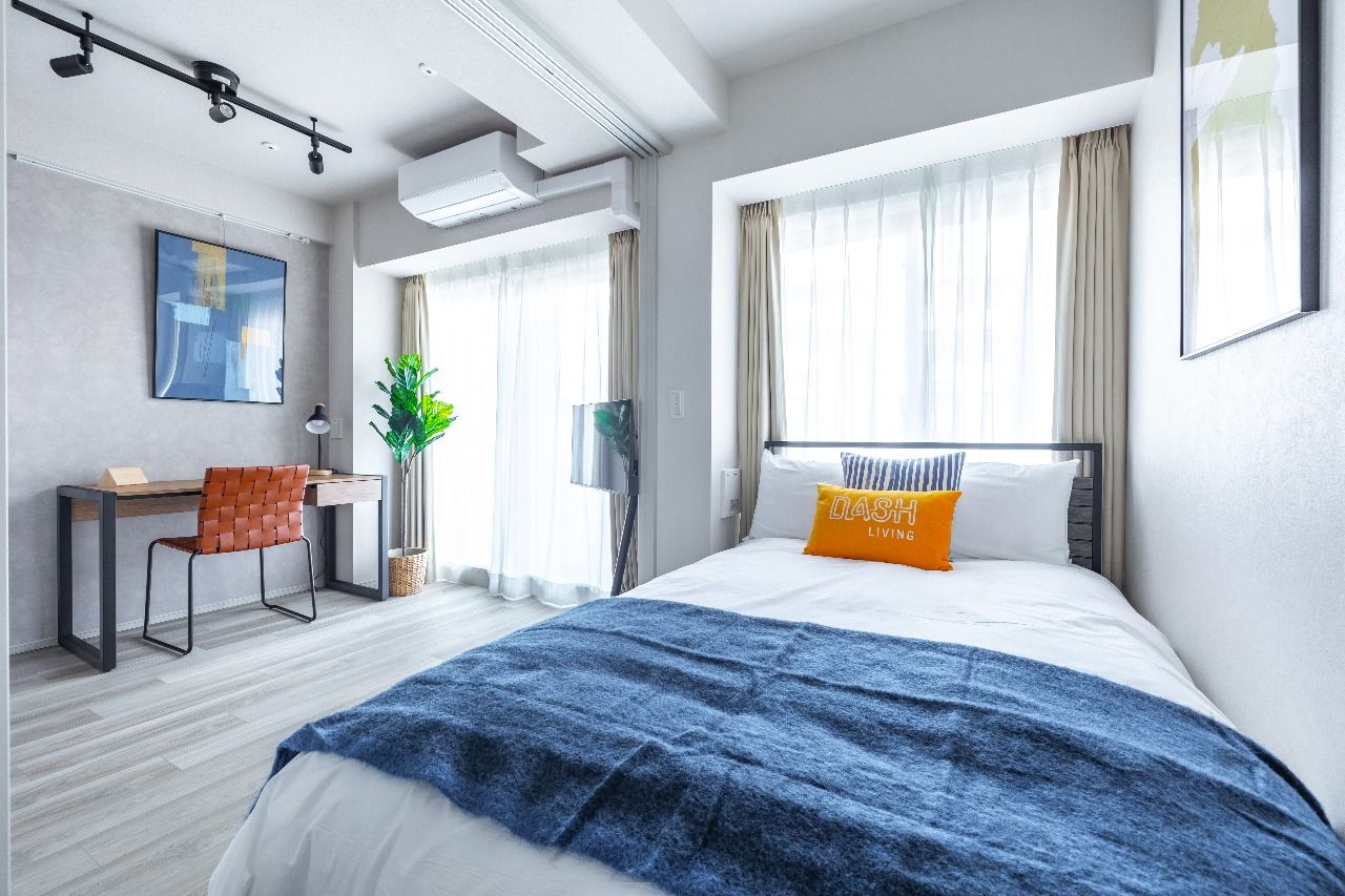 Modern bedroom with a neatly made bed, wooden desk, plant, and large window.