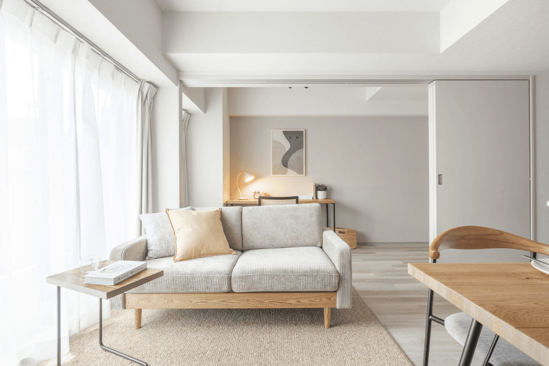 Bright minimalist living room with a light sofa, sheer curtains, and a desk in the background.