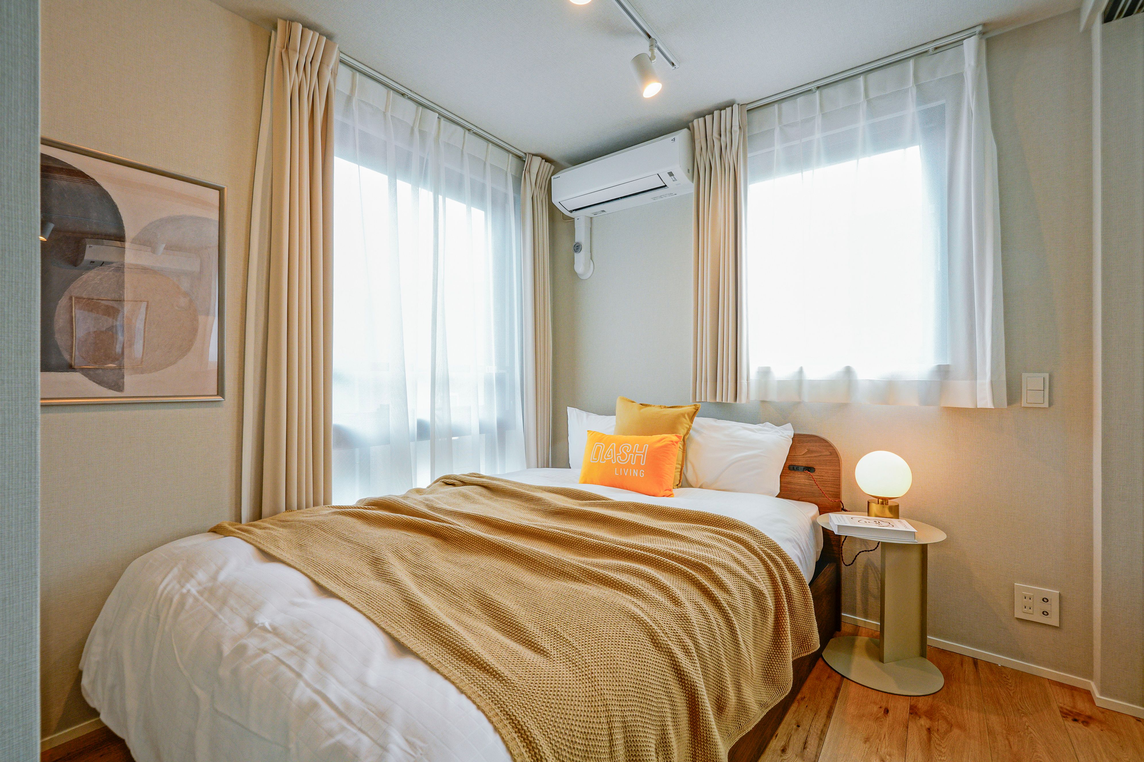 Cozy bedroom with a bed, white sheets, mustard blanket, and two bright windows.