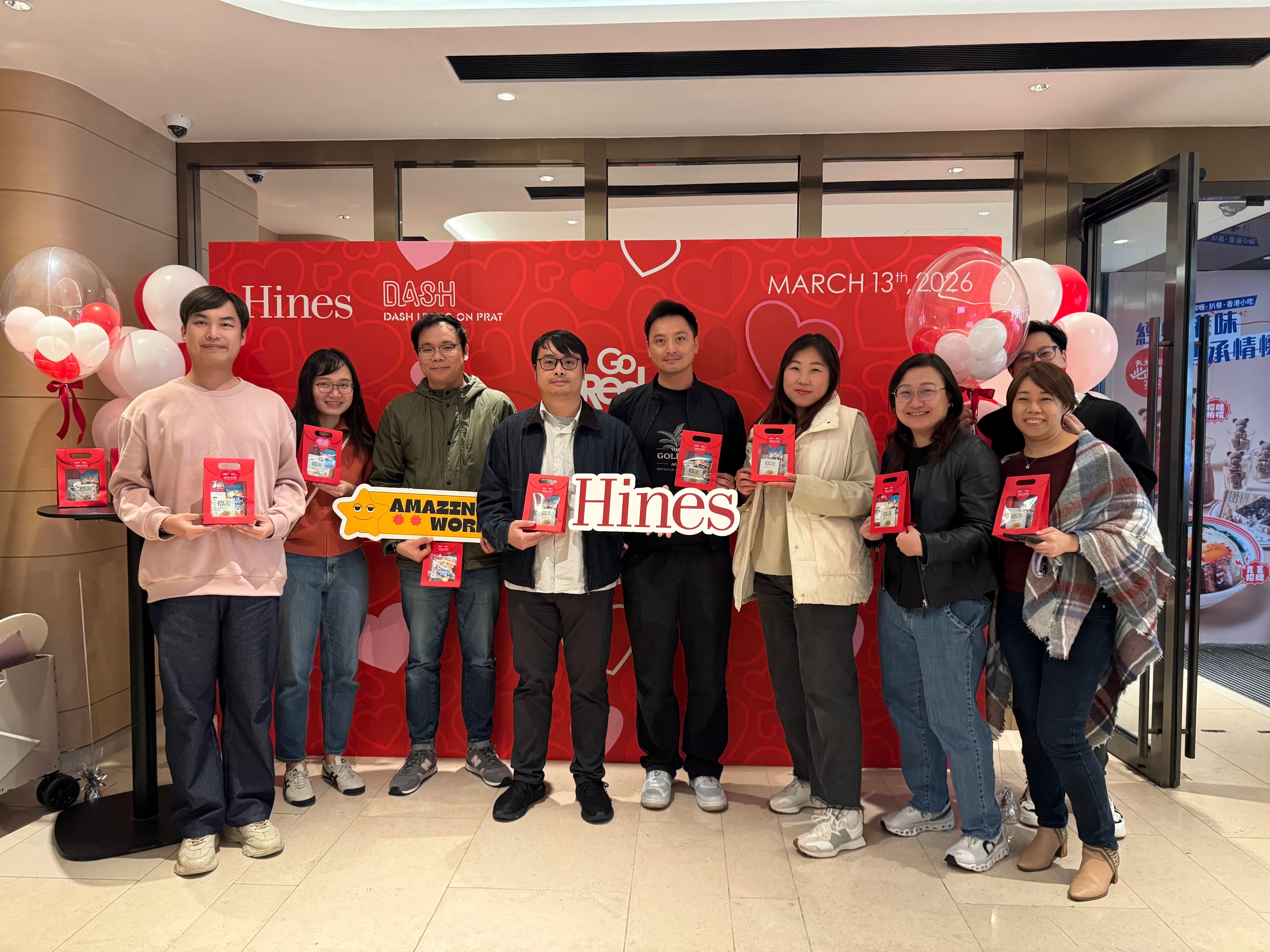 Nine people holding red items at a Hines event with a red backdrop and balloons.