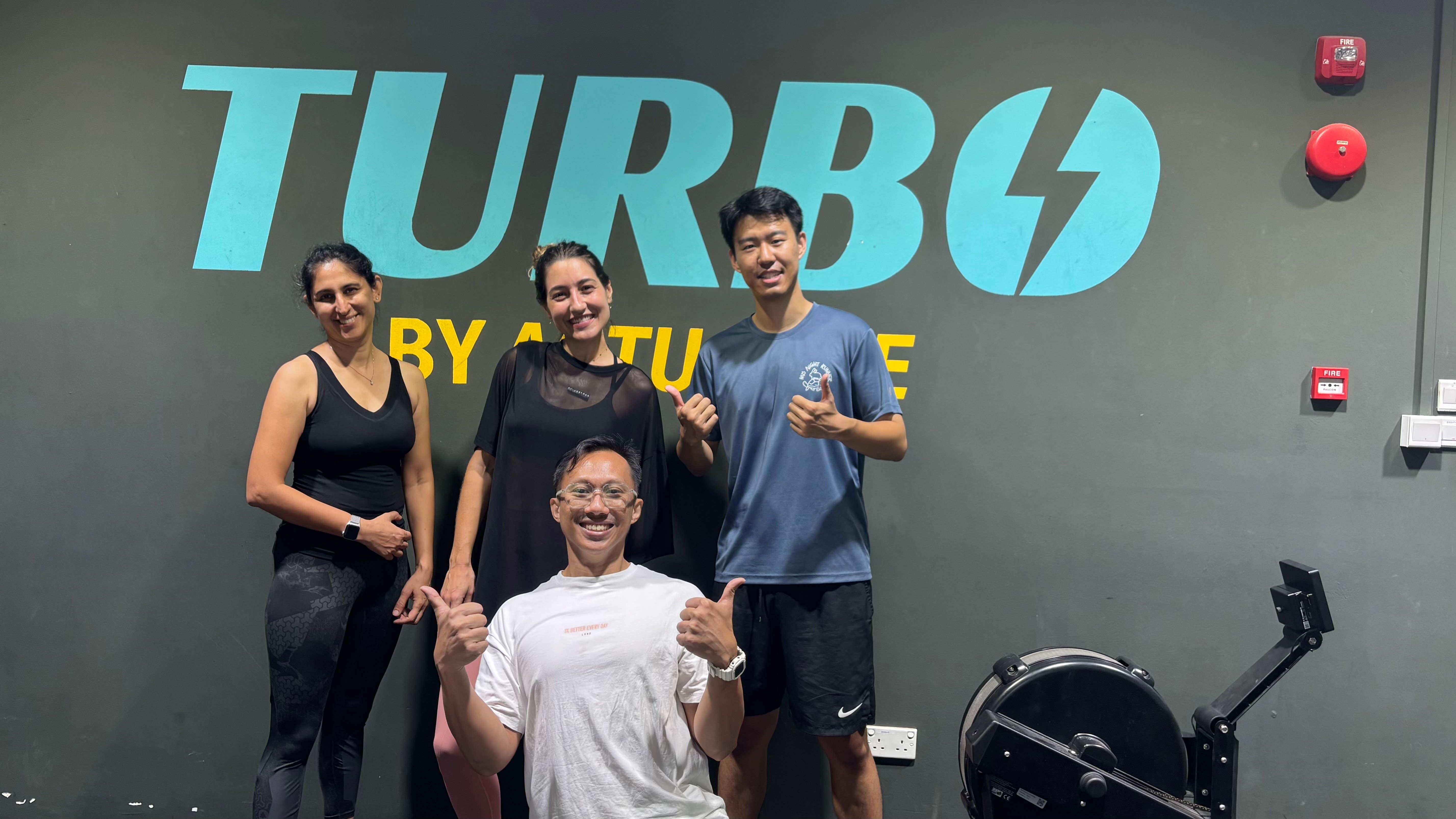 Four smiling people give thumbs-up in a gym, with a prominent 'TURBO' sign.