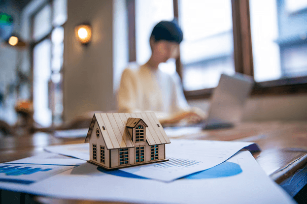 Wooden house model on financial papers, with a blurred person working on a laptop.