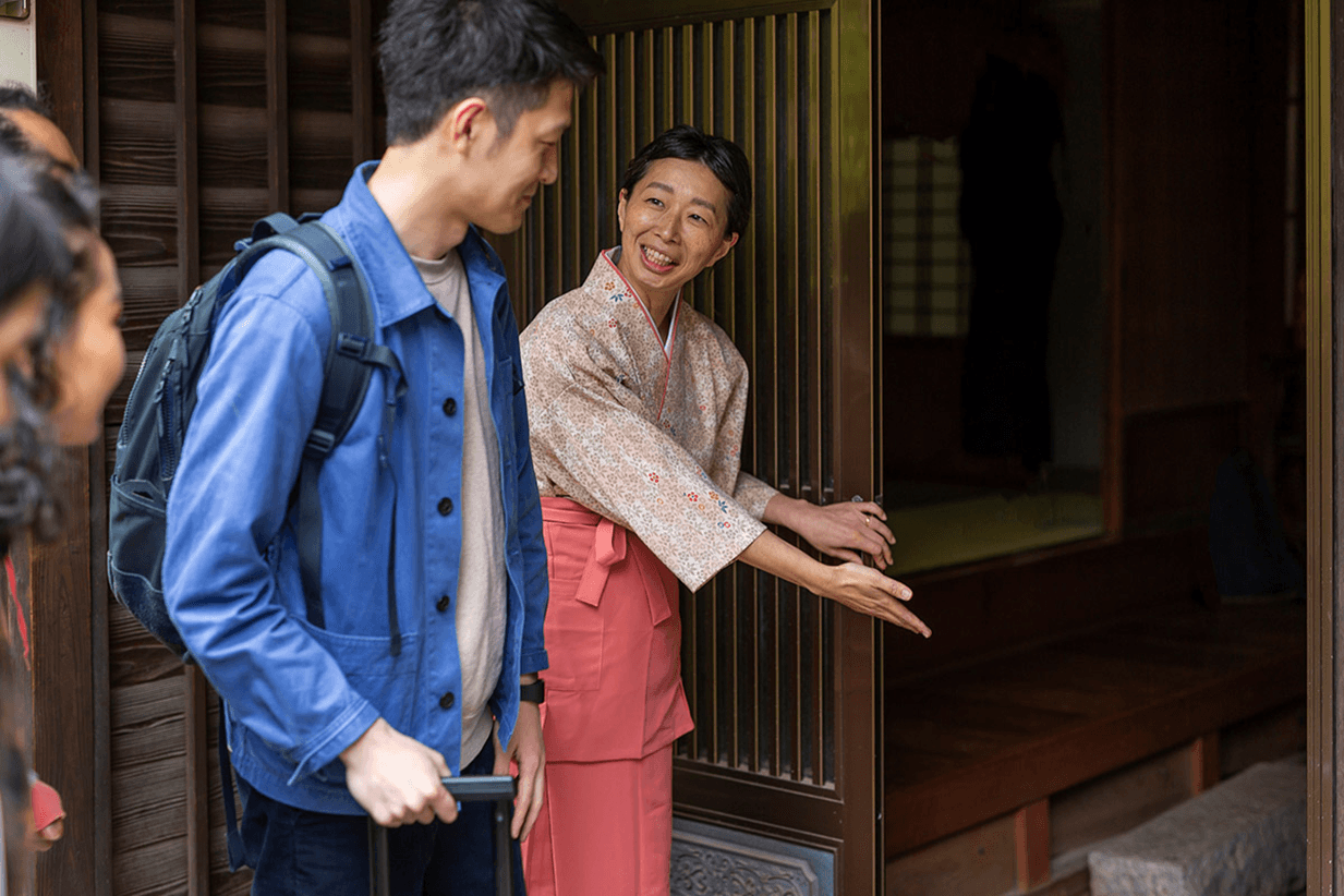 A smiling Japanese woman in traditional attire welcomes a man into a historic building.
