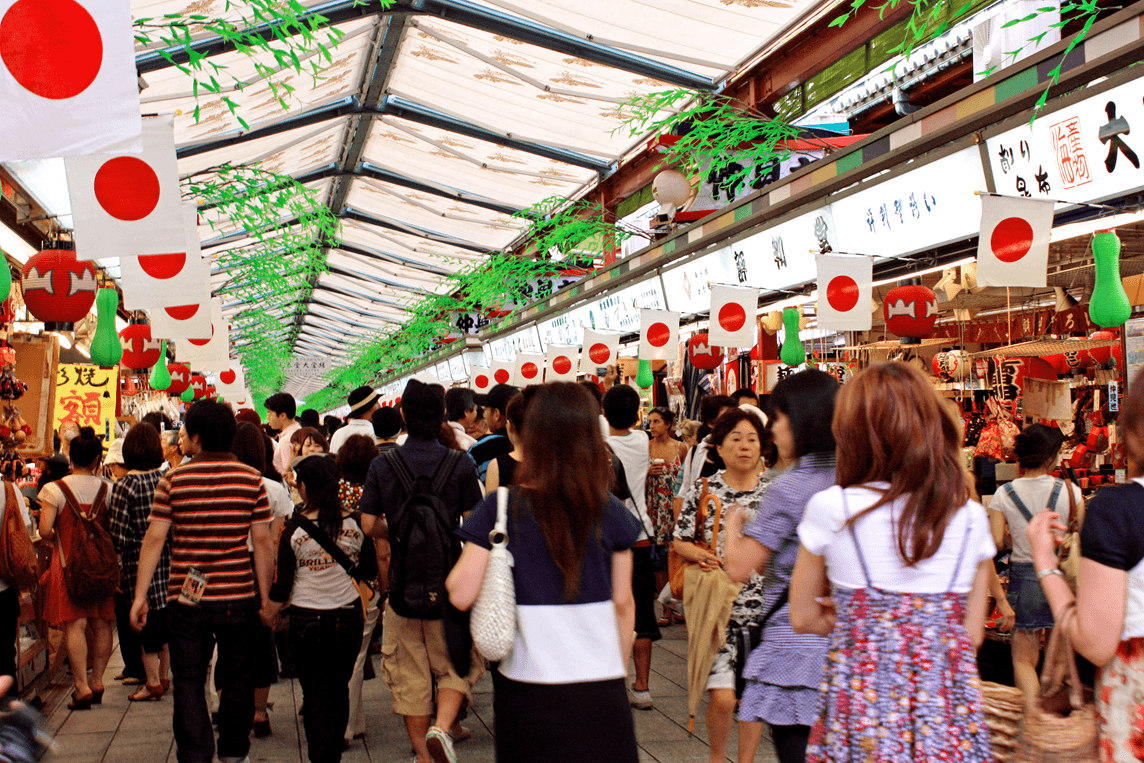 Crowded Japanese market street with people walking under a covered walkway adorned with flags.