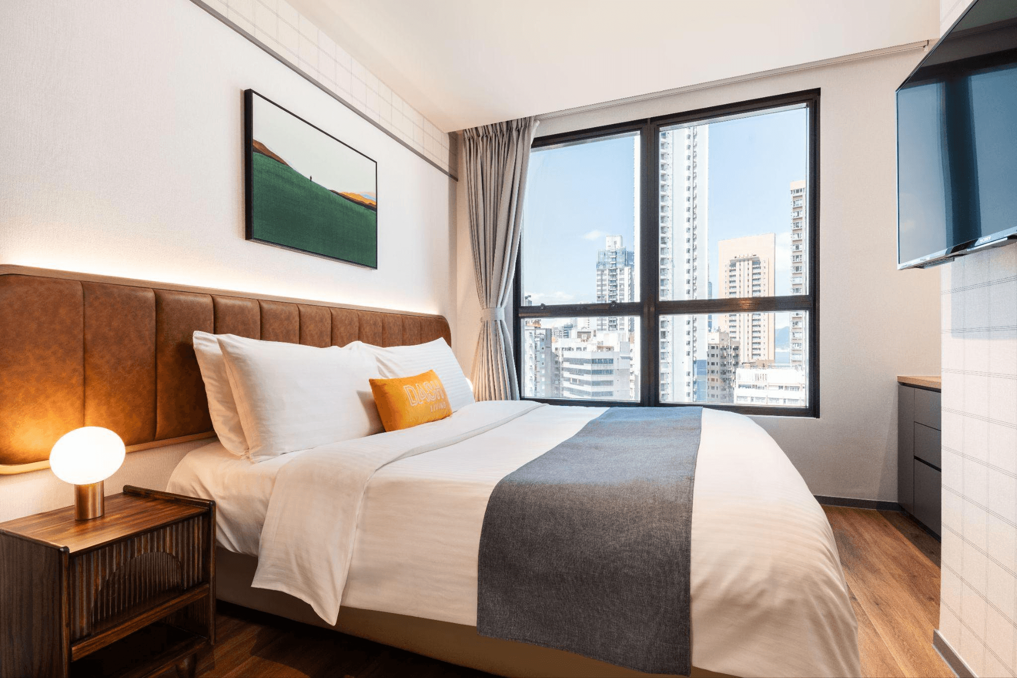 Modern hotel bedroom with a large bed, brown headboard, and a city view window.