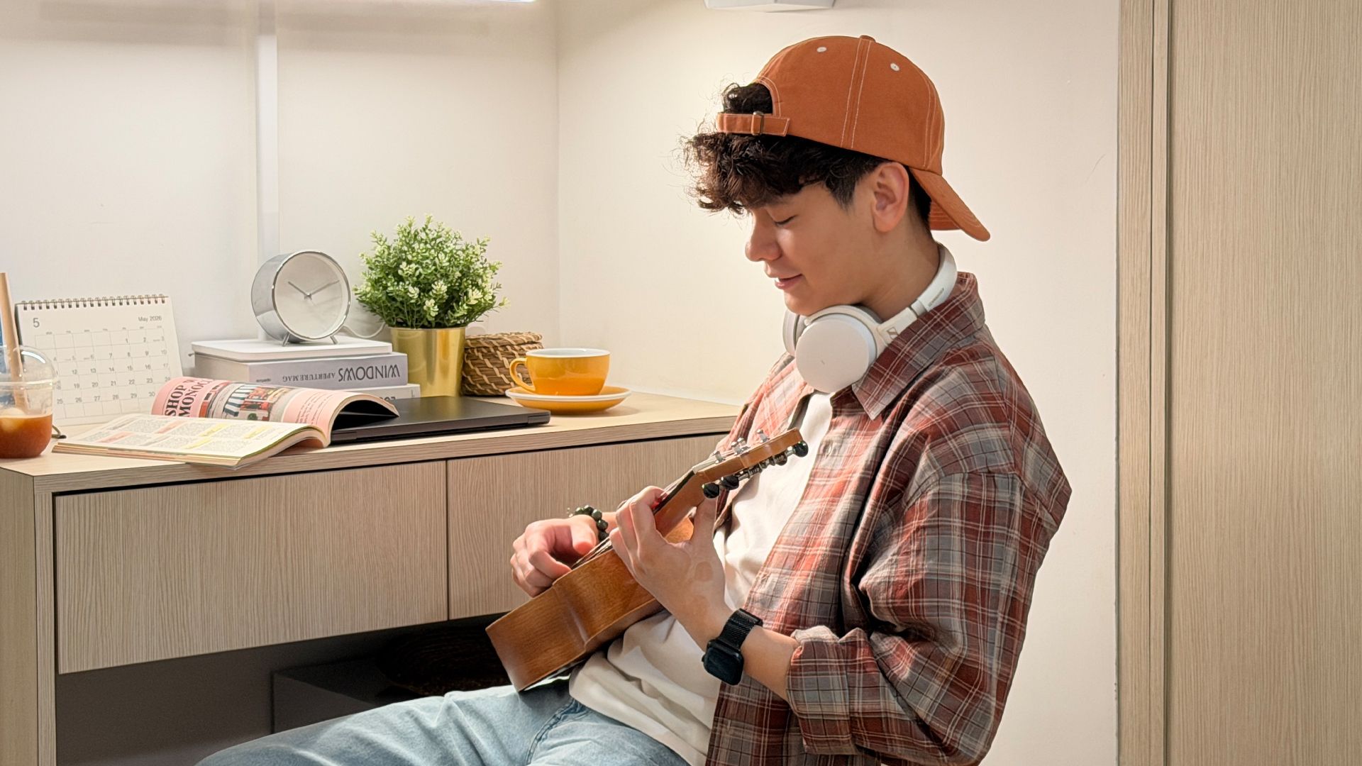 Young man with an orange cap playing a ukulele at a light wooden desk indoors.