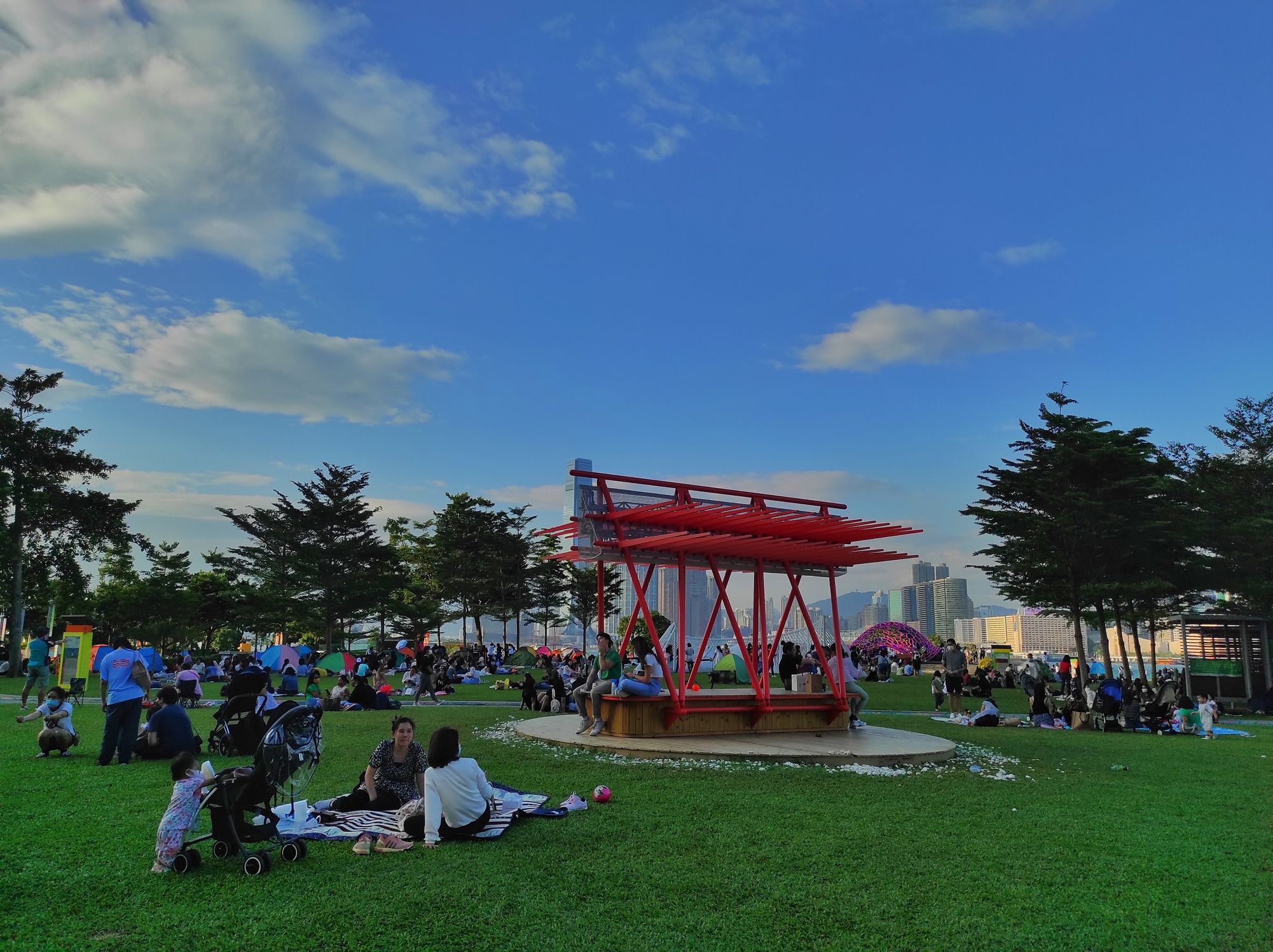 People picnic on green grass around a red pagoda-like structure in a lively park.