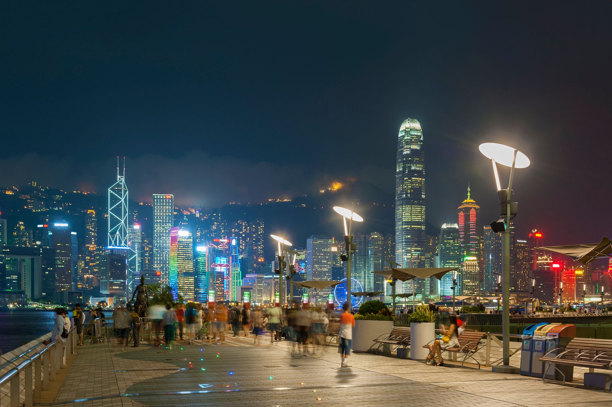 Vibrant city skyline at night, seen from a bustling waterfront promenade with blurred figures.