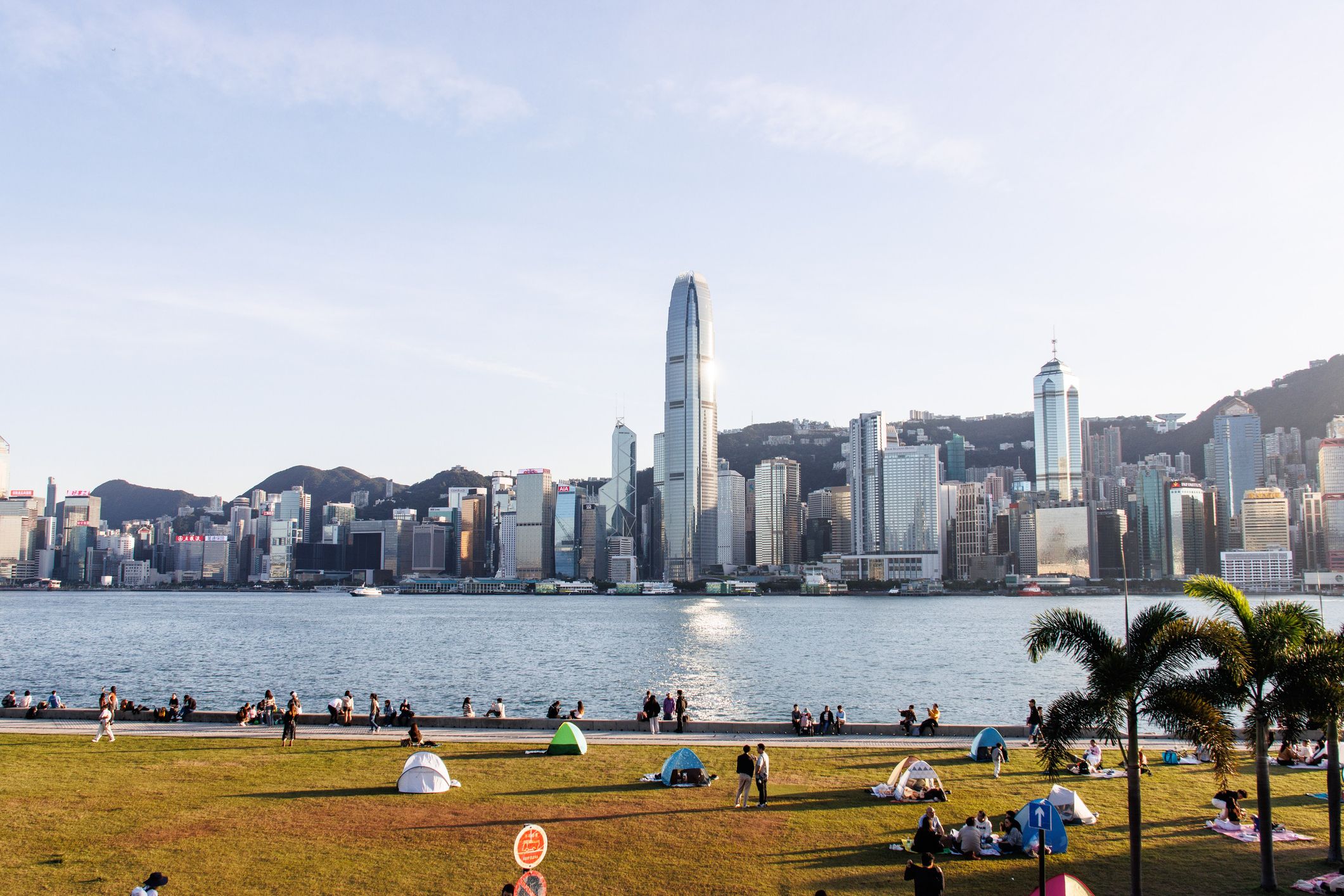 Hong Kong skyline across a harbor, with people relaxing in a sunny green park.