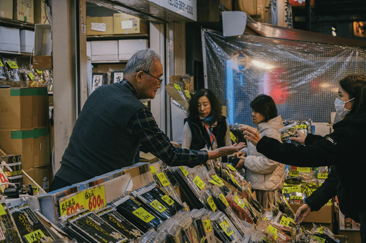 An elderly Japanese vendor sells packaged goods to customers at a busy market stall.