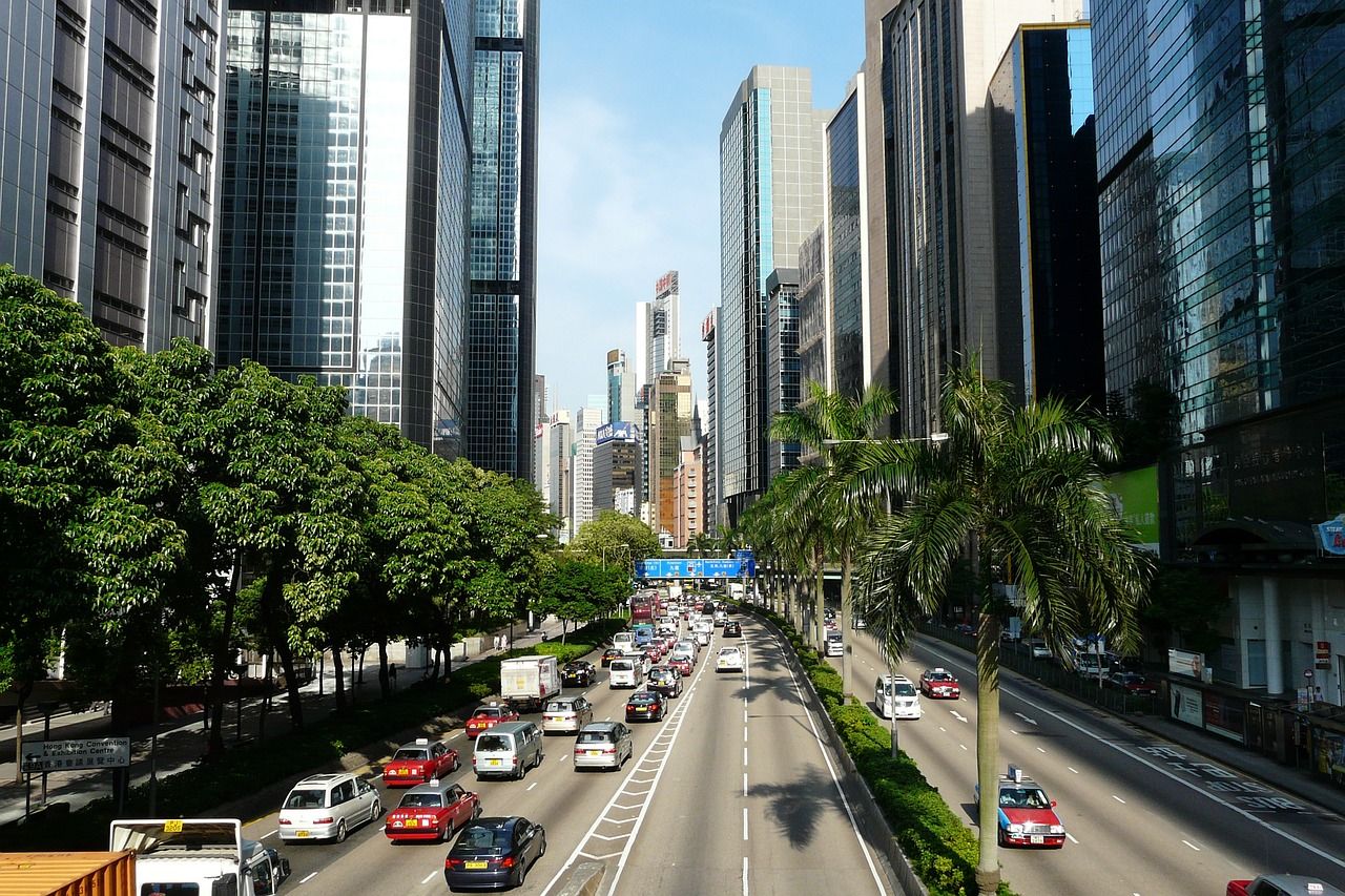 Busy city street with numerous cars, tall skyscrapers, and green trees under a clear sky.