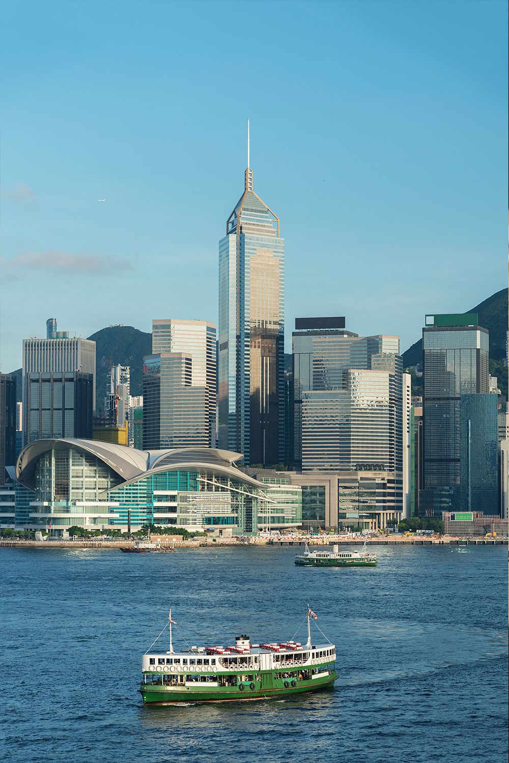 Green ferry crossing water with Hong Kong's towering modern city skyline in the background.