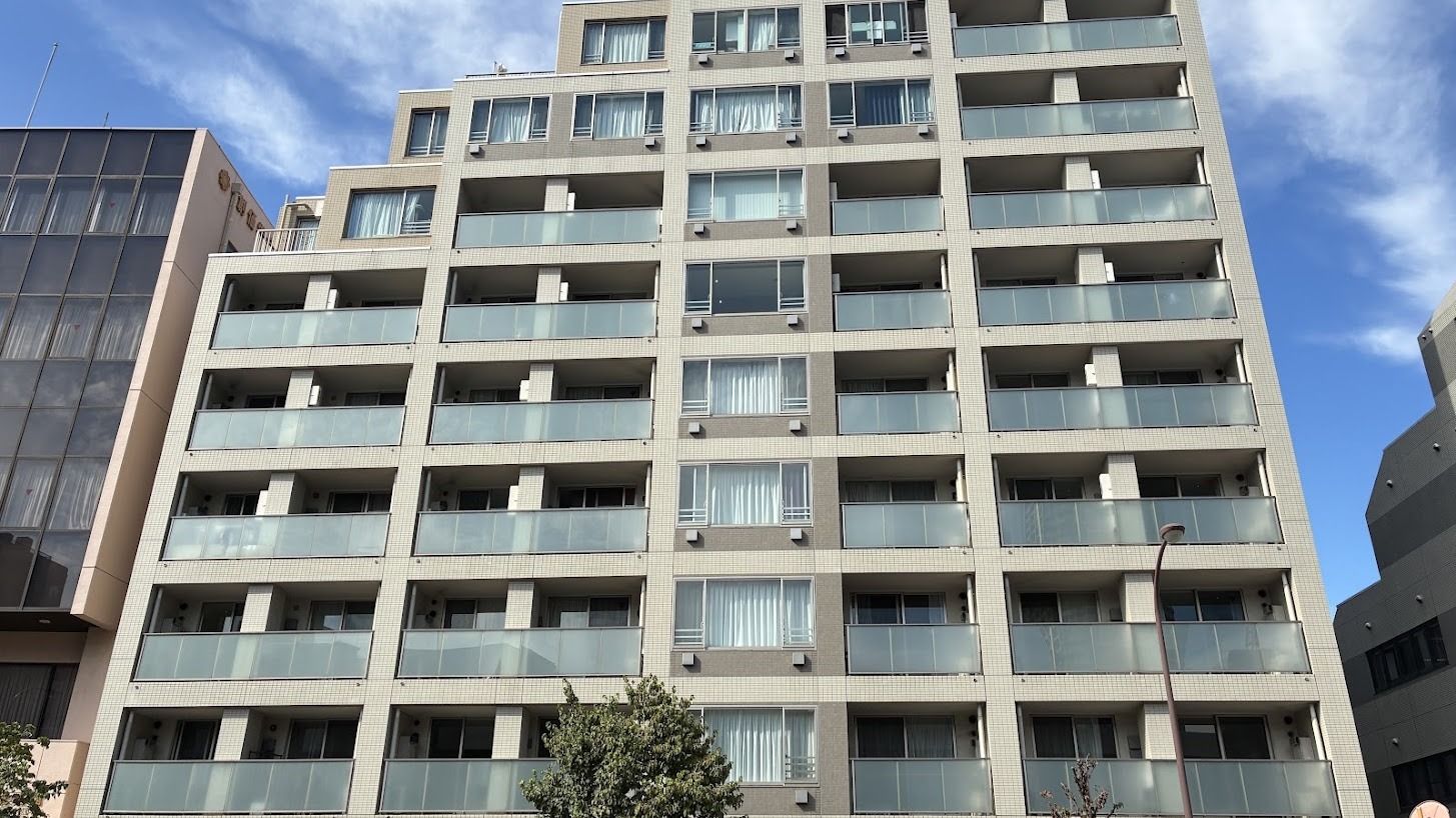 Modern multi-story building with numerous balconies featuring clear glass railings under a cloudy sky.