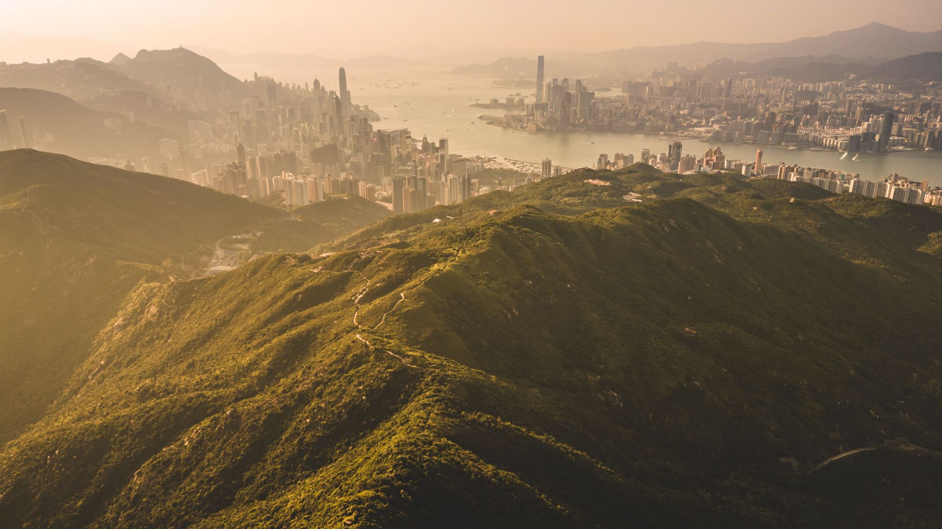 Hazy aerial view of green mountain ridges overlooking a vibrant city skyline by the water.