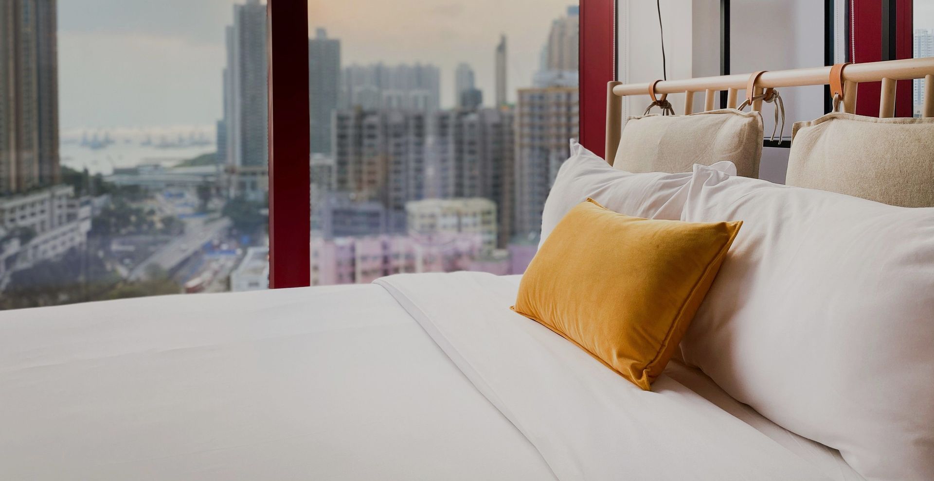 A modern bedroom with a white bed and yellow pillow, overlooking a city skyline. || 'hero section image'