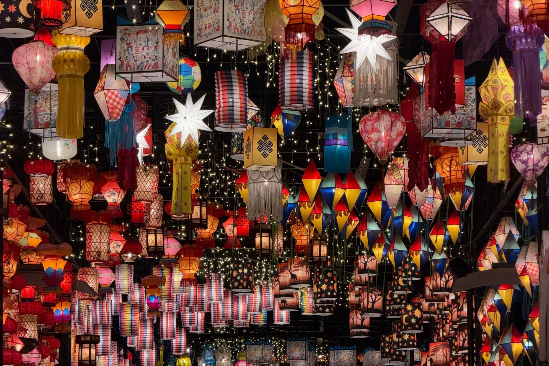 Numerous colorful, illuminated paper lanterns of various shapes hang closely together, creating a festive display.