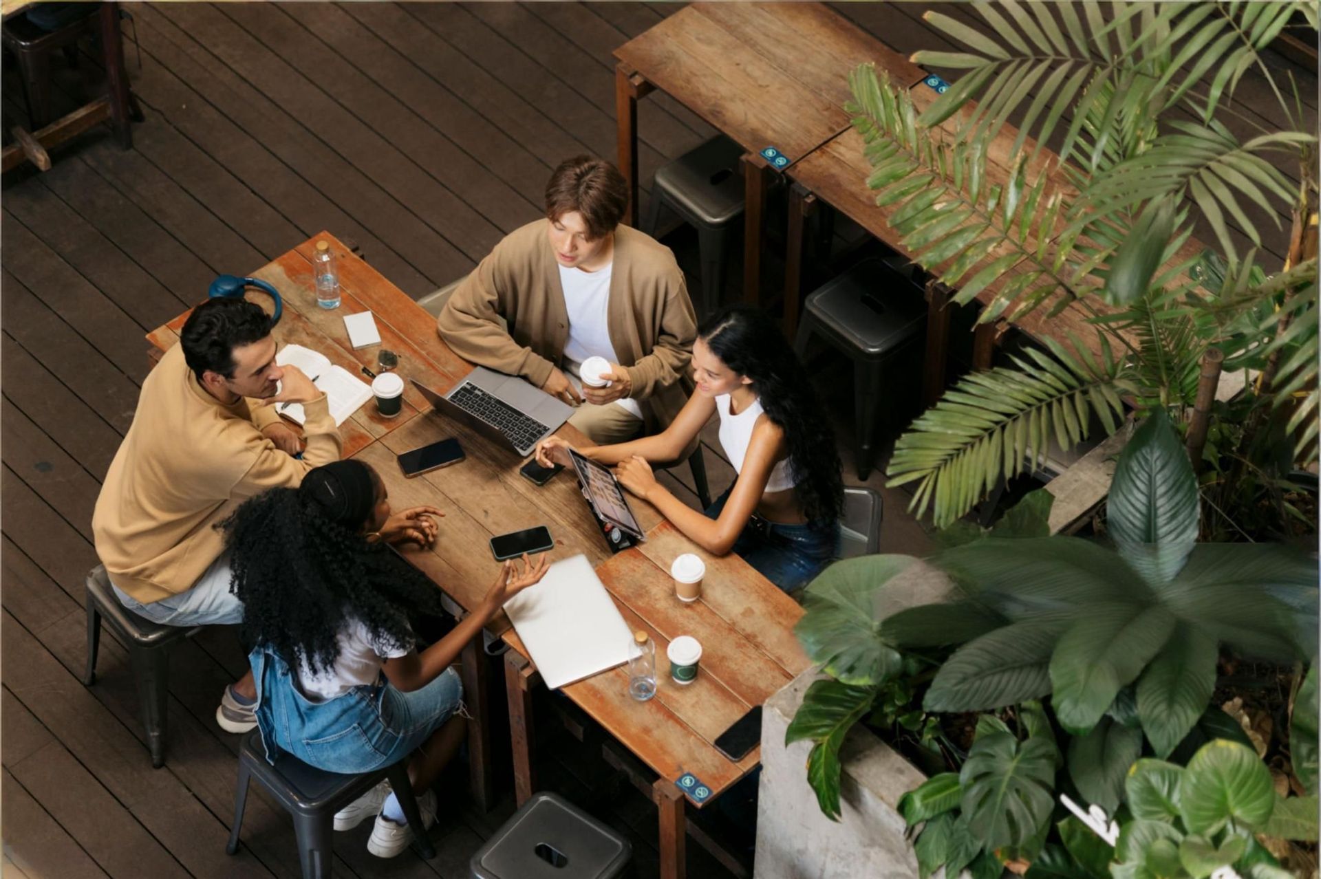 Four people collaborate around a wooden table in a cafe, surrounded by lush green plants.